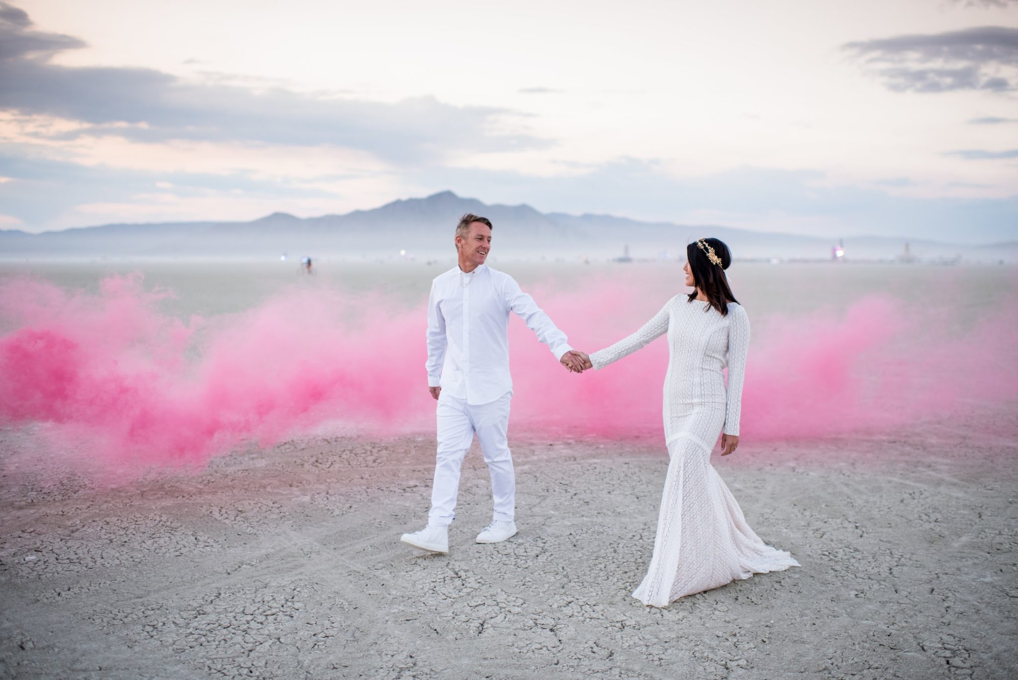 A man and a woman in white wedding clothes hold hands and look at each other as they walk in front of a pink smoke cloud in the desert.