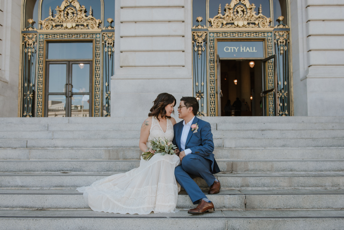 A woman in an off-white wedding dress sits on the steps in front of city hall next to a woman with short hair in a blue formal suit, and they look at each other lovingly.
