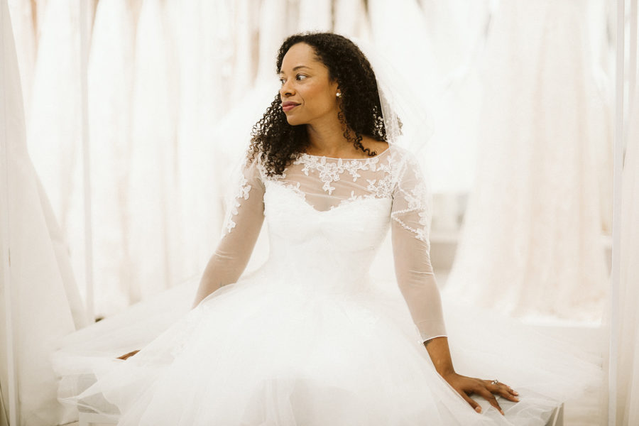 A woman sits in a bridal fitting room wearing a long sleeve wedding gown. She looks to one side, thoughtfully.