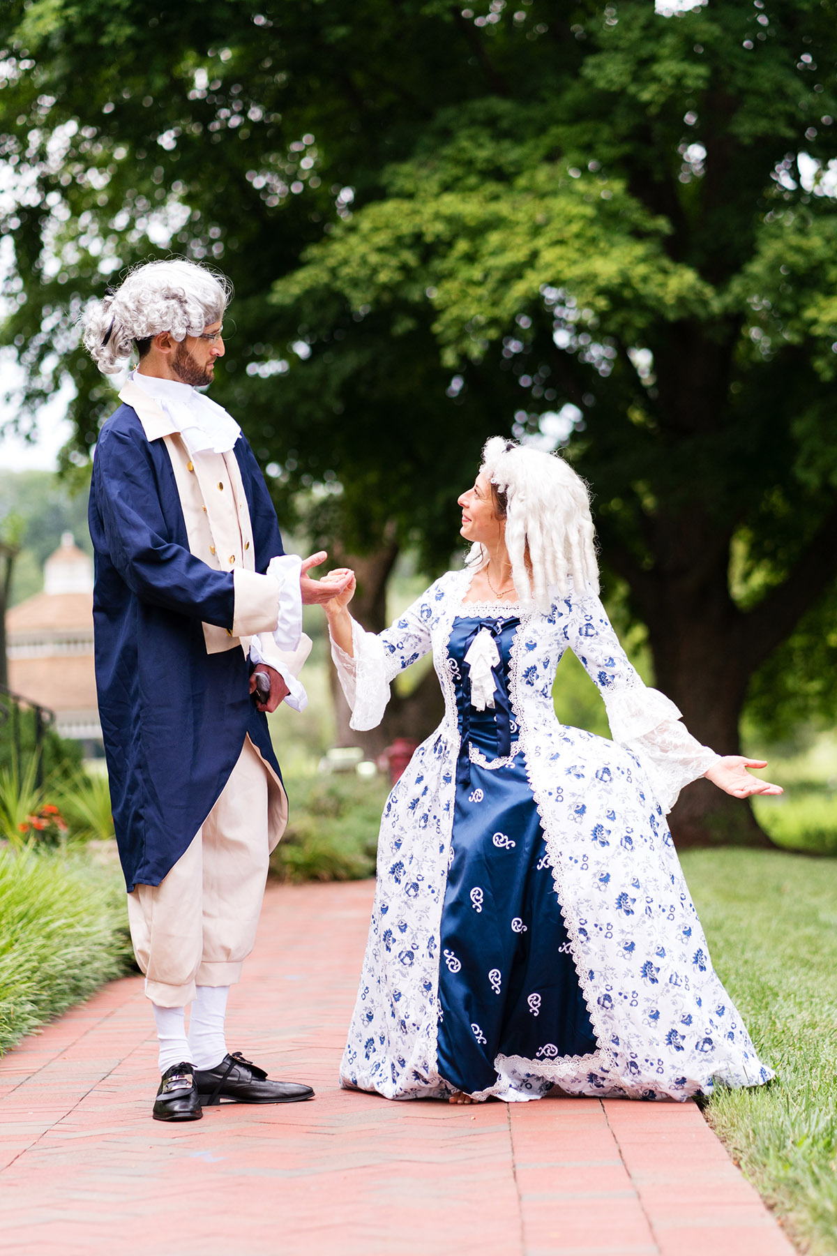 A man and woman wearing 18th century European fancy dress and white wigs hold hands and look at one another with amused expressions as the woman curtseys. 