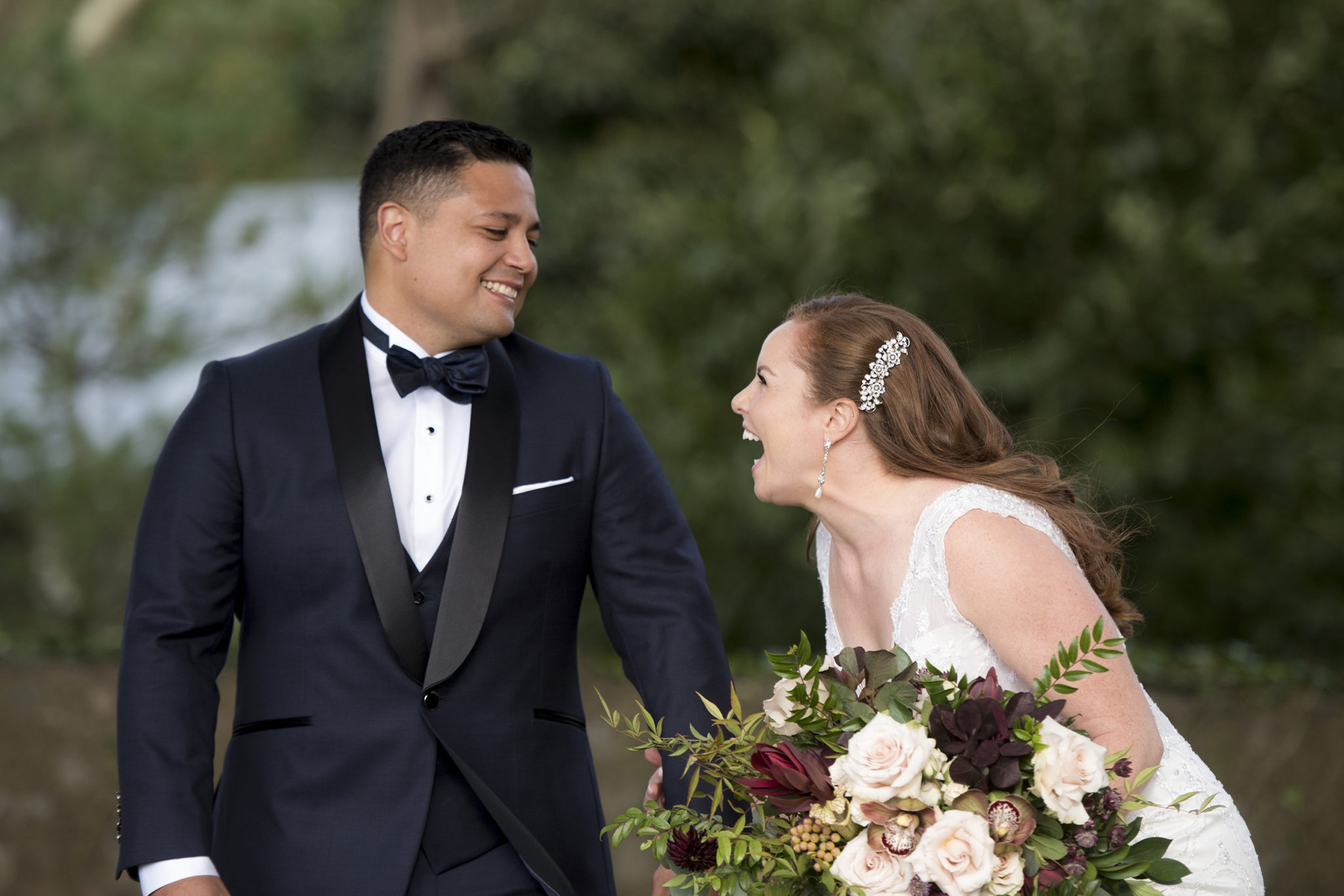 A woman in a white wedding dress and long auburn hair laughs while looking at and holding hands with a groom in a black tux who smiles back