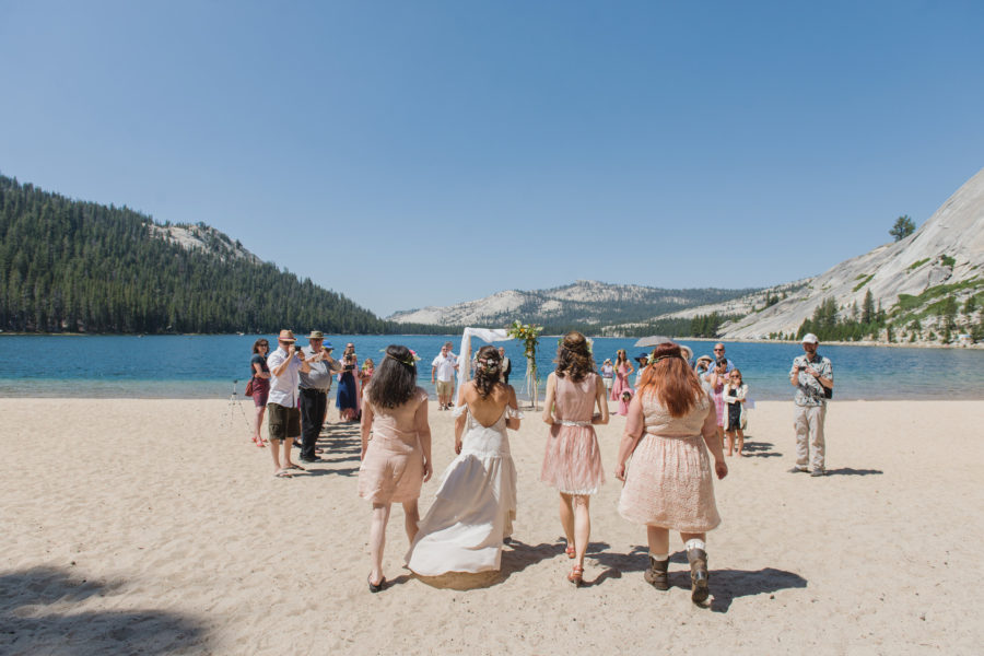 A wedding party in white and pink processes toward a wedding arch on a sandy beach overlooking water and mountains at a destination wedding
