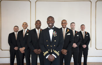 A Marine groom in uniform jacket and bow tie is flanked by six groomsmen in tuxes. All are smiling.