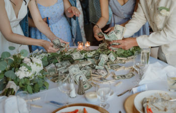 A bride, groom, and two women in blue dresses gathered around a pile of cash on a wedding reception table after dinner