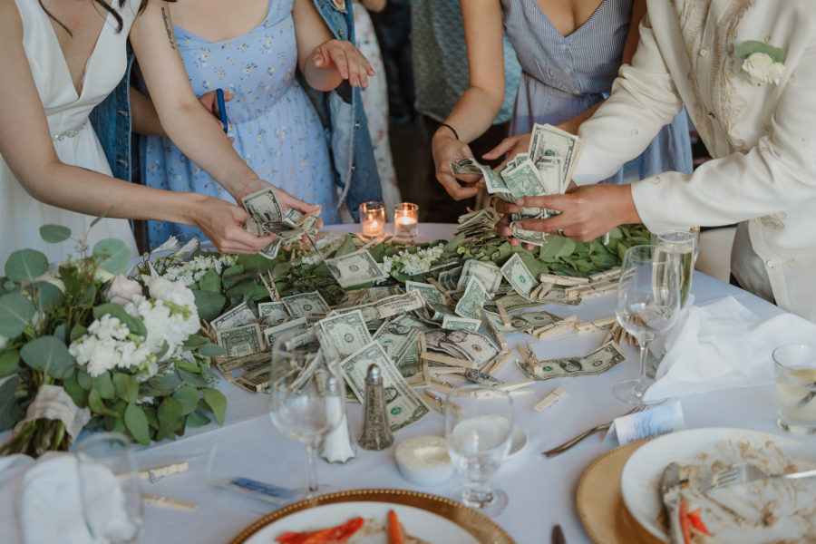 A bride, groom, and two women in blue dresses gathered around a pile of cash on a wedding reception table after dinner