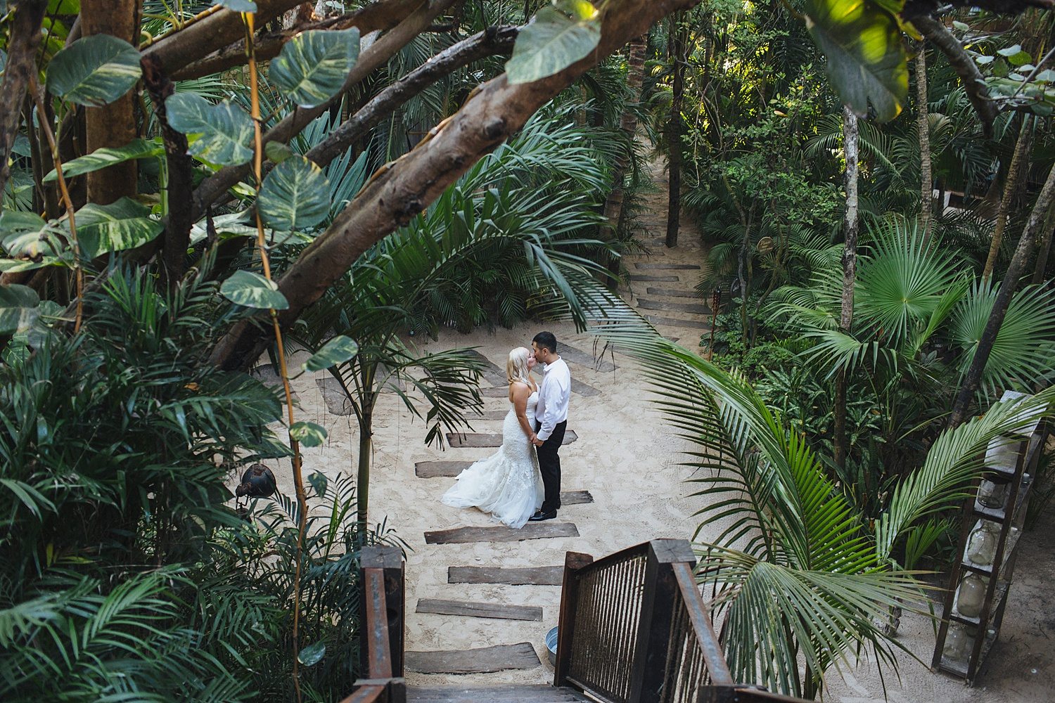 A couple in a wedding dress and shirt sleeves from a black tux stand amidst lush green tropical plants on a sandy path at their destination wedding
