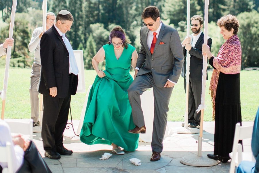 A woman in a green wedding dress and a man in a grey suit smash a glass each at the end of their Jewish wedding ceremony, surrounded by loved ones