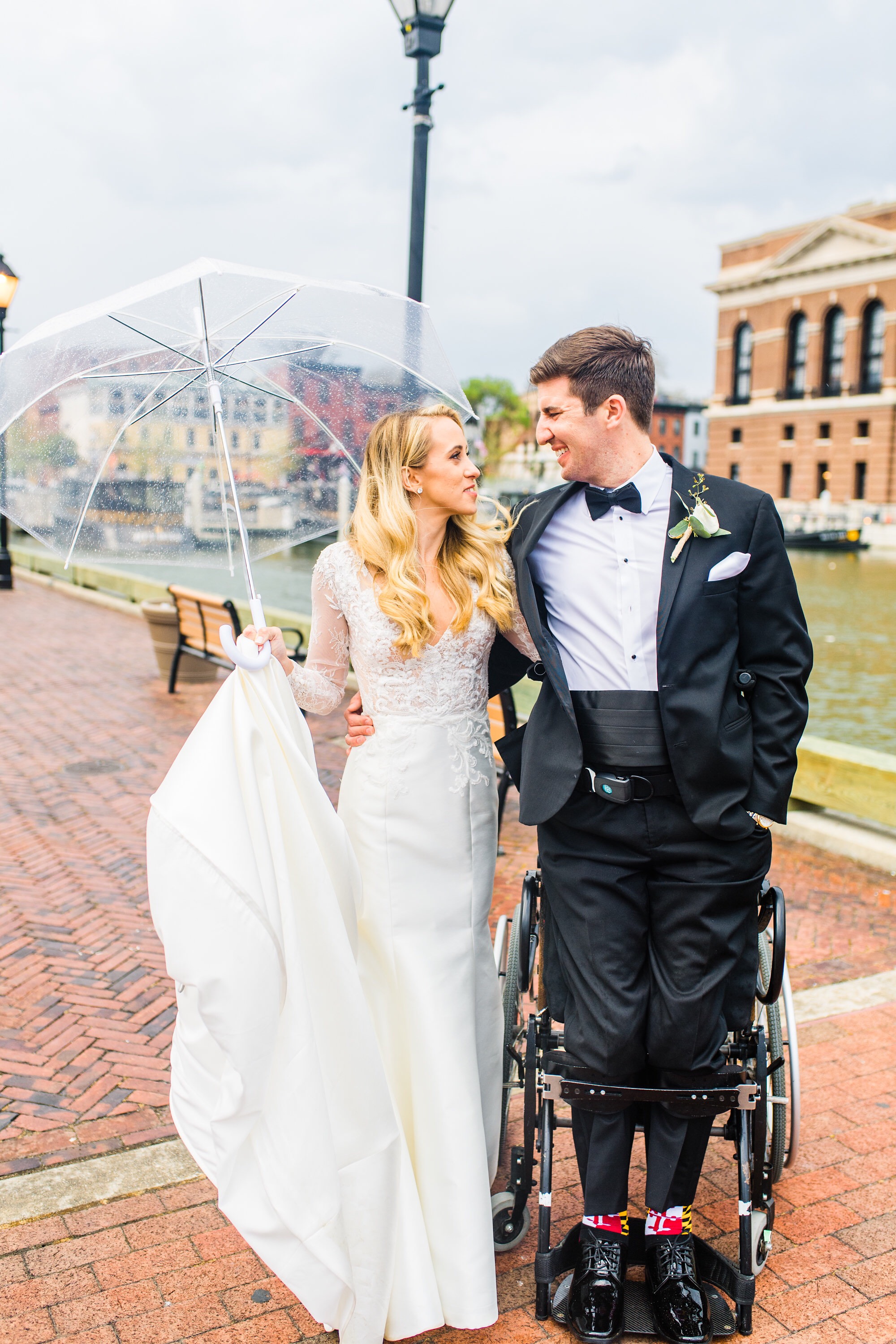 a bride and groom standing on a brick walkway. The bride holds a clear wedding umbrella for rain and the groom is standing up in a wheelchair supported by leg braces