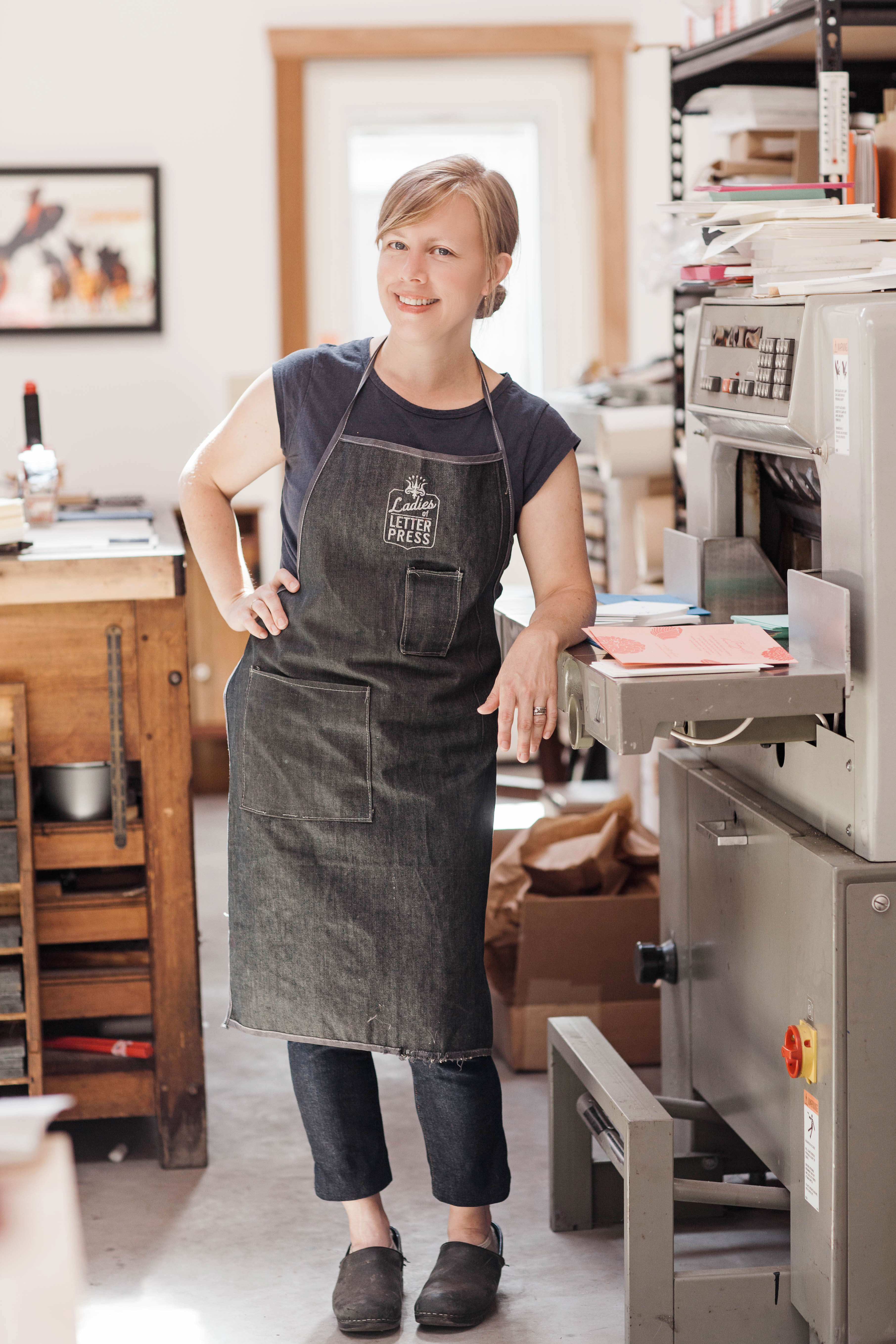 portrait of Kseniya Thomas of Thomas Printers wearing a Ladies Who Letterpress apron and standing at her letterpress 