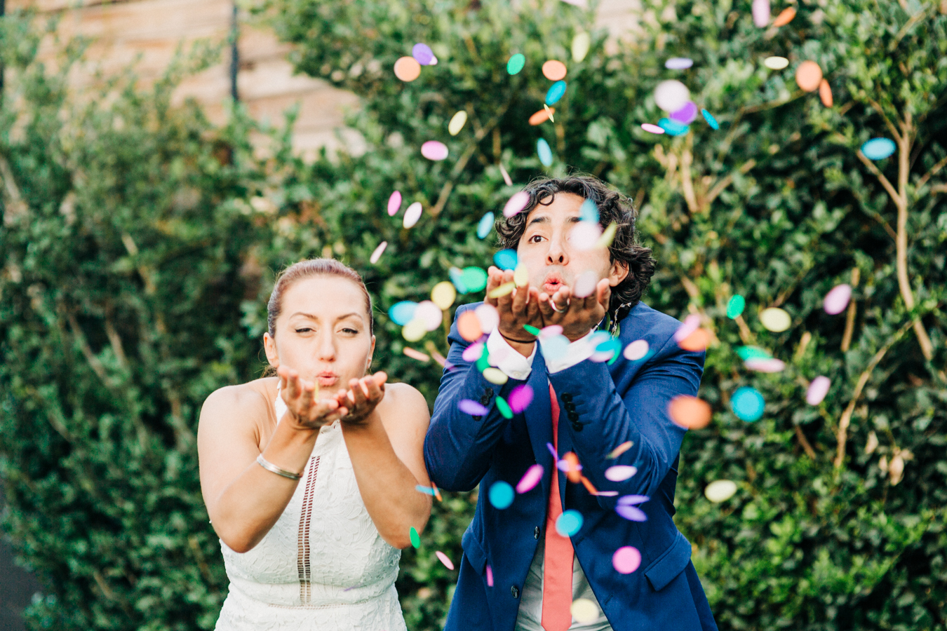 bride and groom standing in front of a hedge holding their hands up in front of them and blowing confetti into the air in a photo by Laura Ford