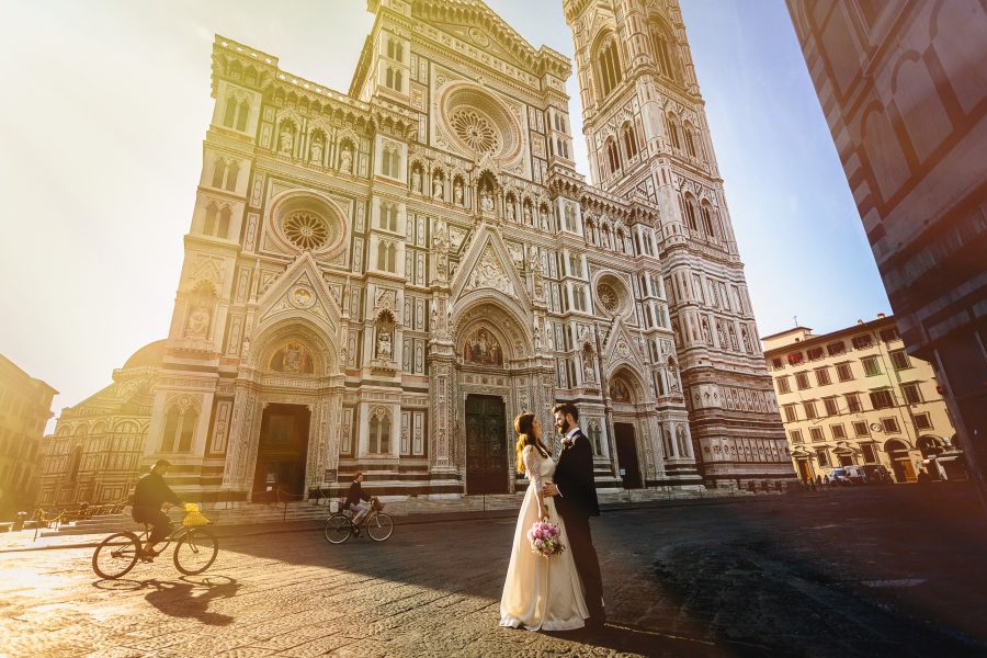 A couple in a wedding dress and tux stand in front of a cathedral smiling at each other at a destination wedding in Italy