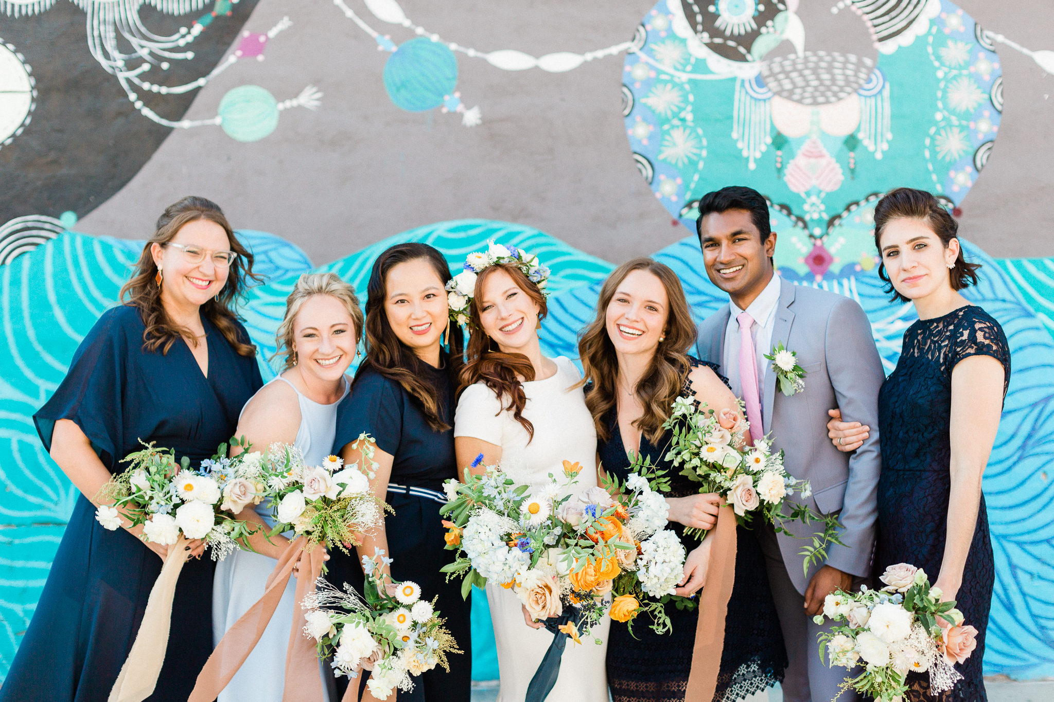 bridal party five woman and one man stand around the bride and face the camera smiling in front of a wall mural in a photo by Laura Ford