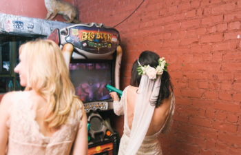 A woman in a wedding dress and veil plays a shooting arcade game, aiming the plastic weapon at the screen