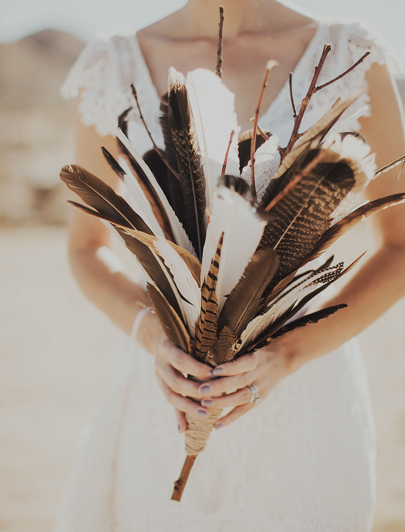 A woman in a white lace wedding dress on a beach holds a bouquet made from white and brown feathers and twigs for her wedding without flowers
