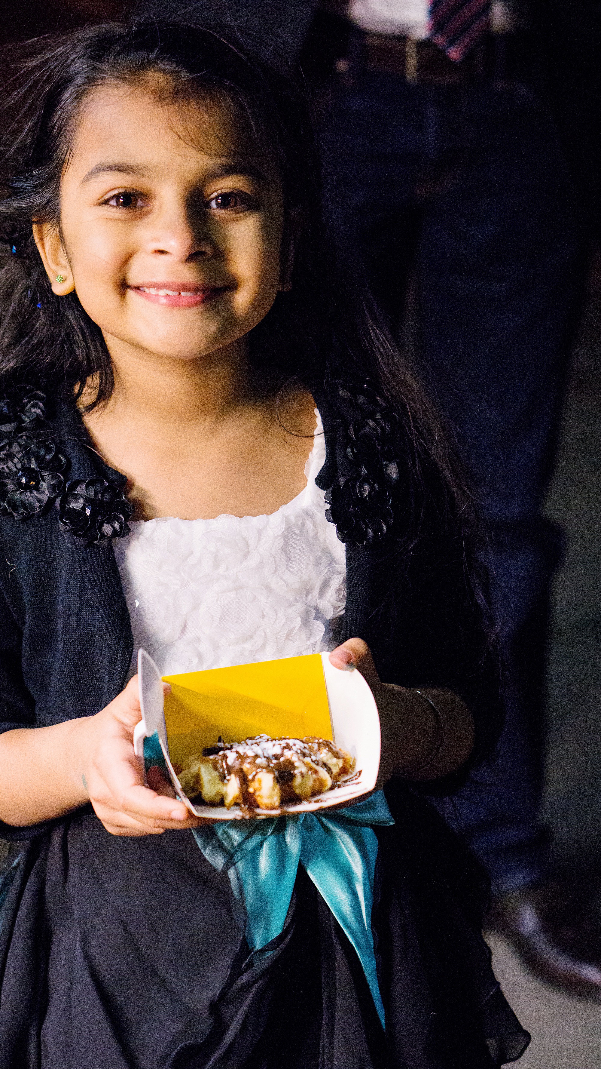 Young girl wearing a flower girl dress with a cardigan is smiling at the camera and holding a Wafels and Dinges waffle in a photo by Kyo Morishima