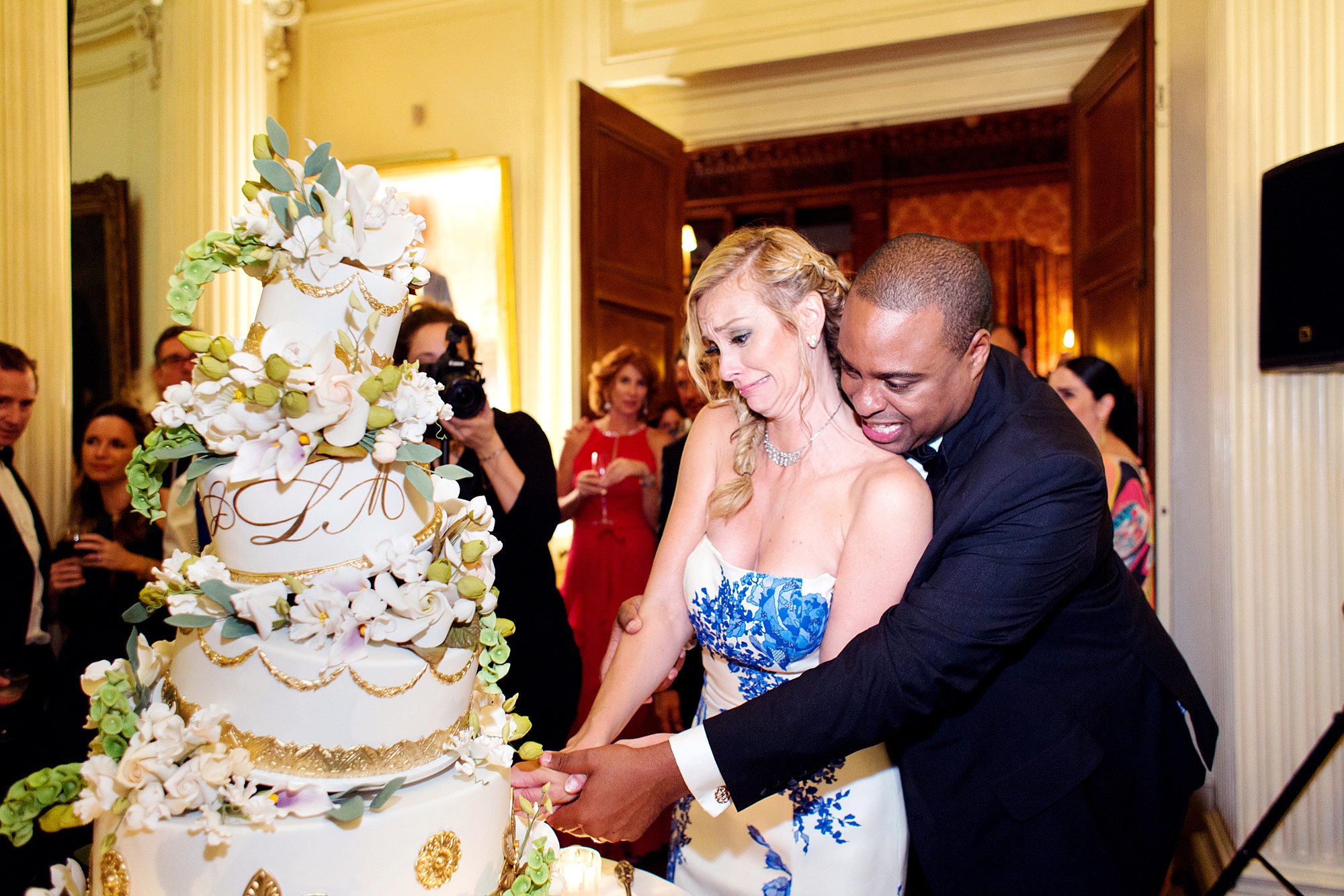 Blond bride wearing a strapless white gown with blue flowers is cutting an enormous elaborately decorated wedding cake, groom stands behind her with his arms around her also helping to cut the cake in a photo by Kyo Morishima