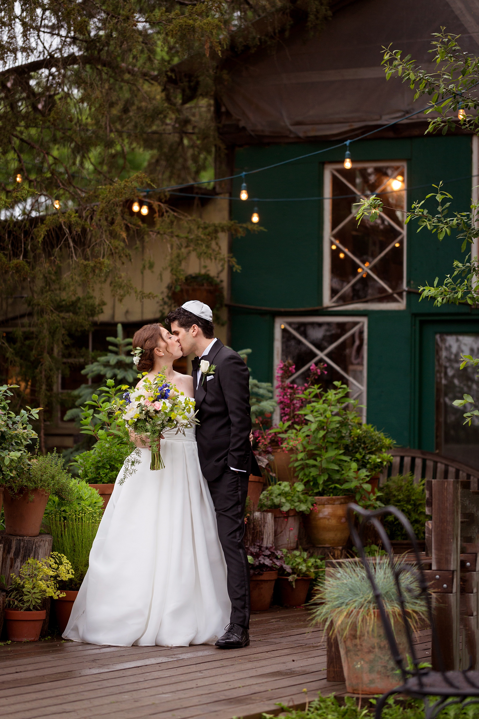 Bride holding large colorful bouquet and groom wearing a yarmulke kiss and stand on an outdoor deck surrounded by plants and lit by bare bulbs in a photo by Kyo Morishima