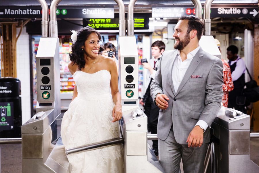 Bride and groom are exiting side-by-side subway turnstiles and looking at each other and grinning in a photo by Kyo Morishima