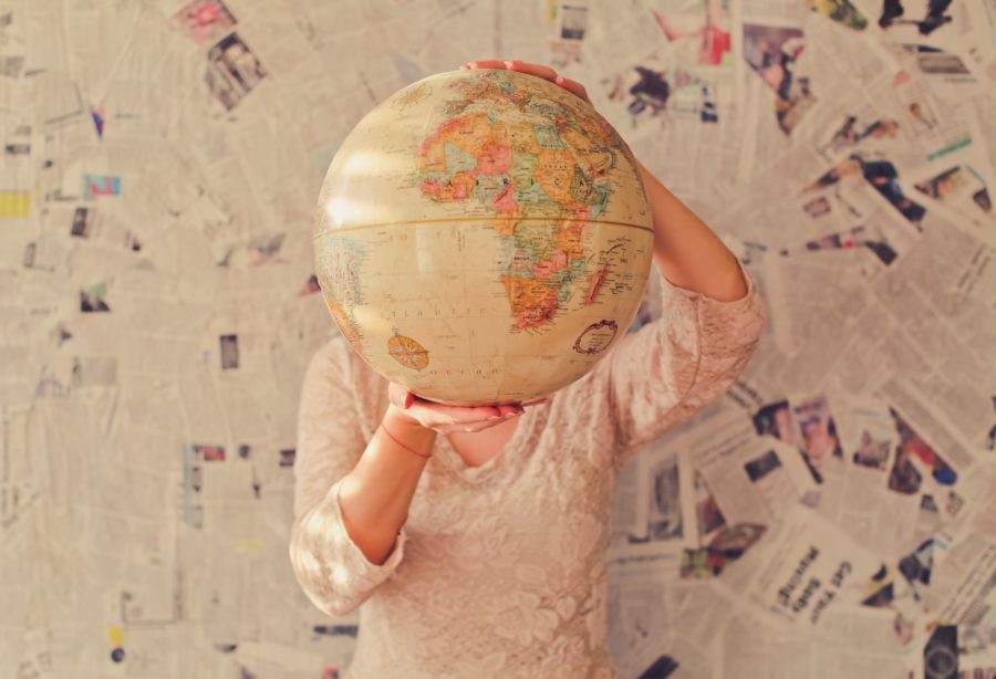 A woman holds a large beige globe in front of her head and torso, while standing in front of a newspaper-covered wall.