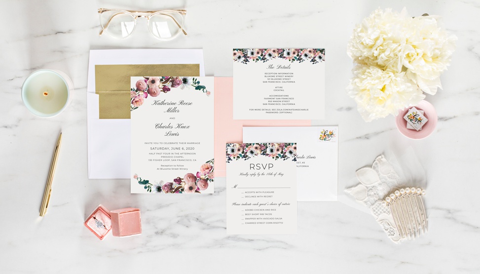 A floral invitation suite from Zola displayed on a marble tabletop with a bunch of white flowers, retro clear frame glasses, a gold pen, and other accessories