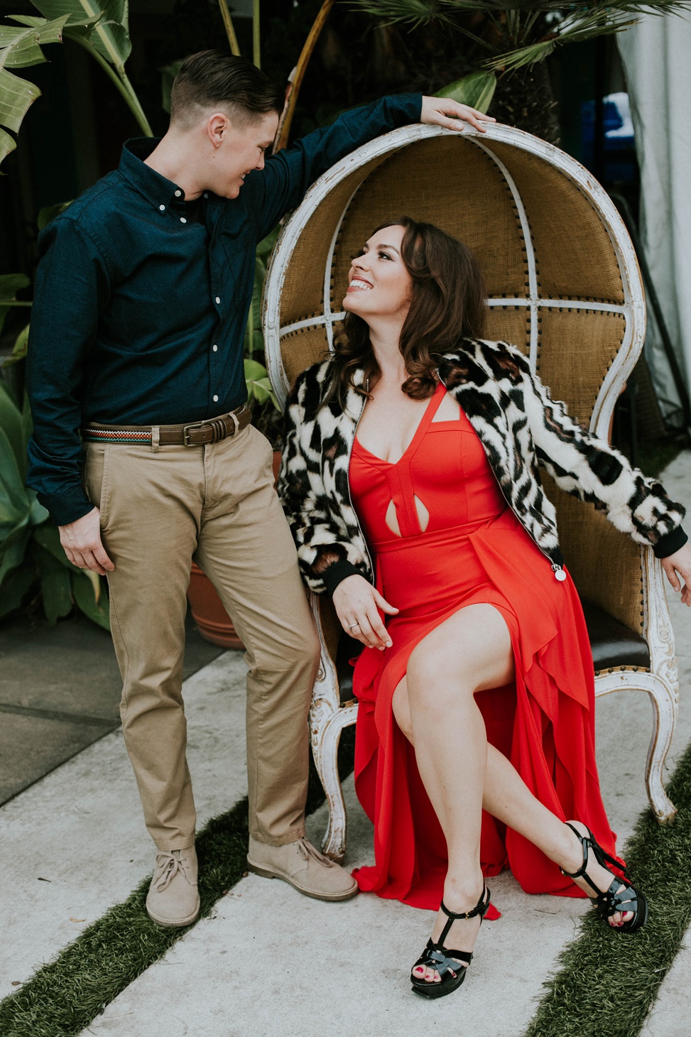 a queer couple pose for an engagement photo wearing a black button down shirt and chinos and a red evening gown with a leopard print faux fur coat