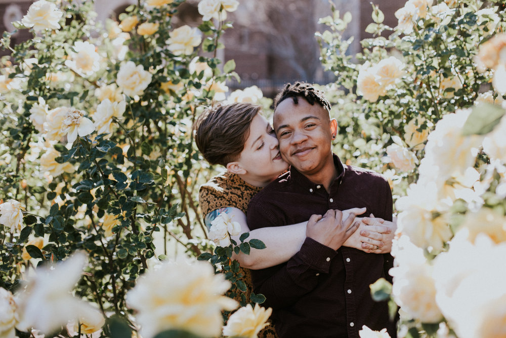 a queer interracial couple poses in an embrace for an engagement photo amidst a rose garden