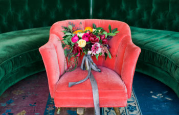 A bouquet of colorful flowers sits on an orange velvet chair in front of a green velvet bench