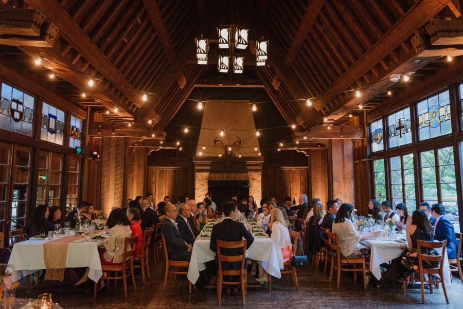 A large party gathers at several family-style tables in a large, high-windowed hall to celebrate an engagement party