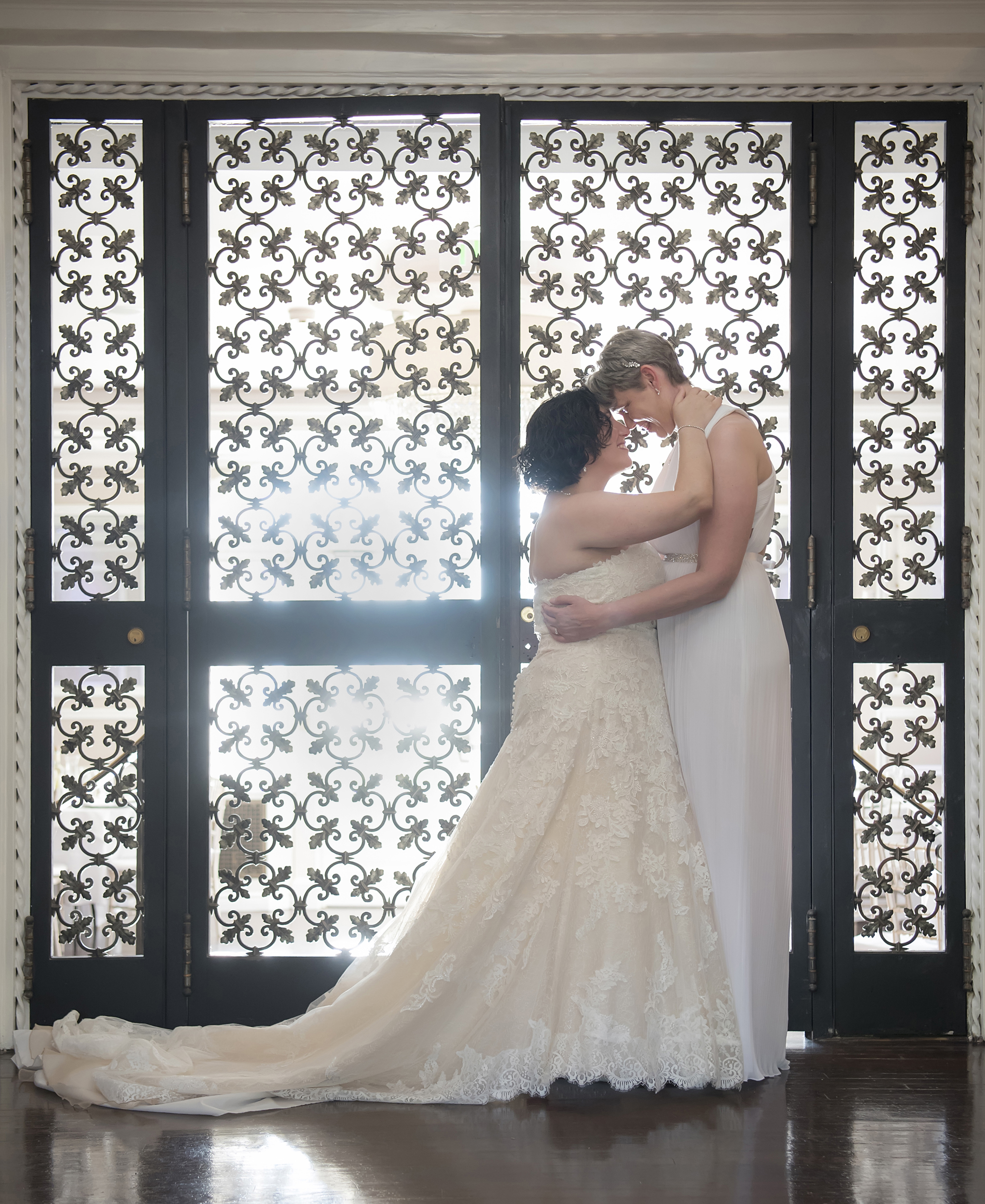 Two brides, one wearing a gown with a train and the other wearing a white jumpsuit stand forehead to forehead in front of a backlit iron screen in a photo by Studio A Images