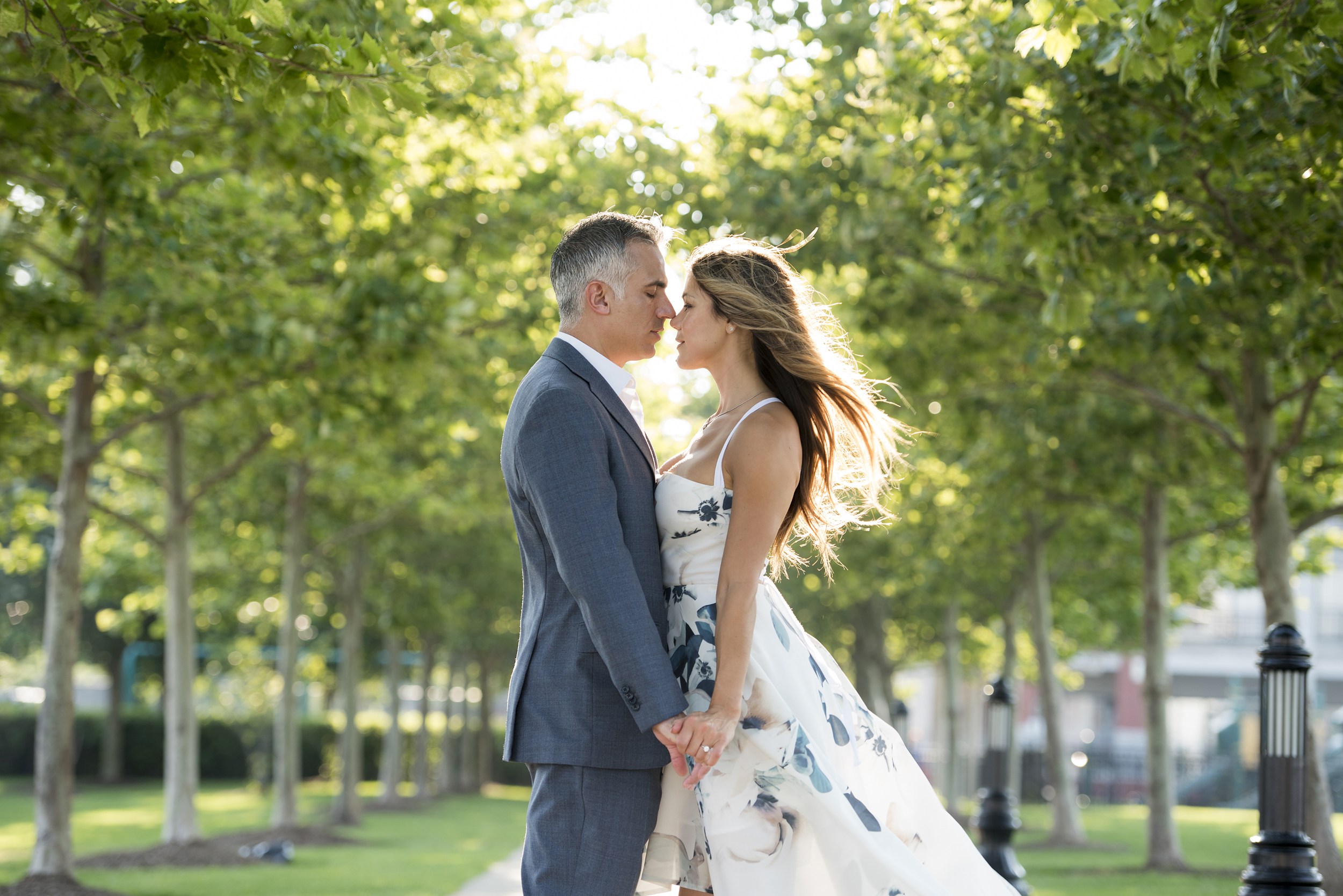 A man in a grey suit and a woman in a white floral dress are about to kiss while standing in a sunlit allee of trees in a photo by Studio A Images