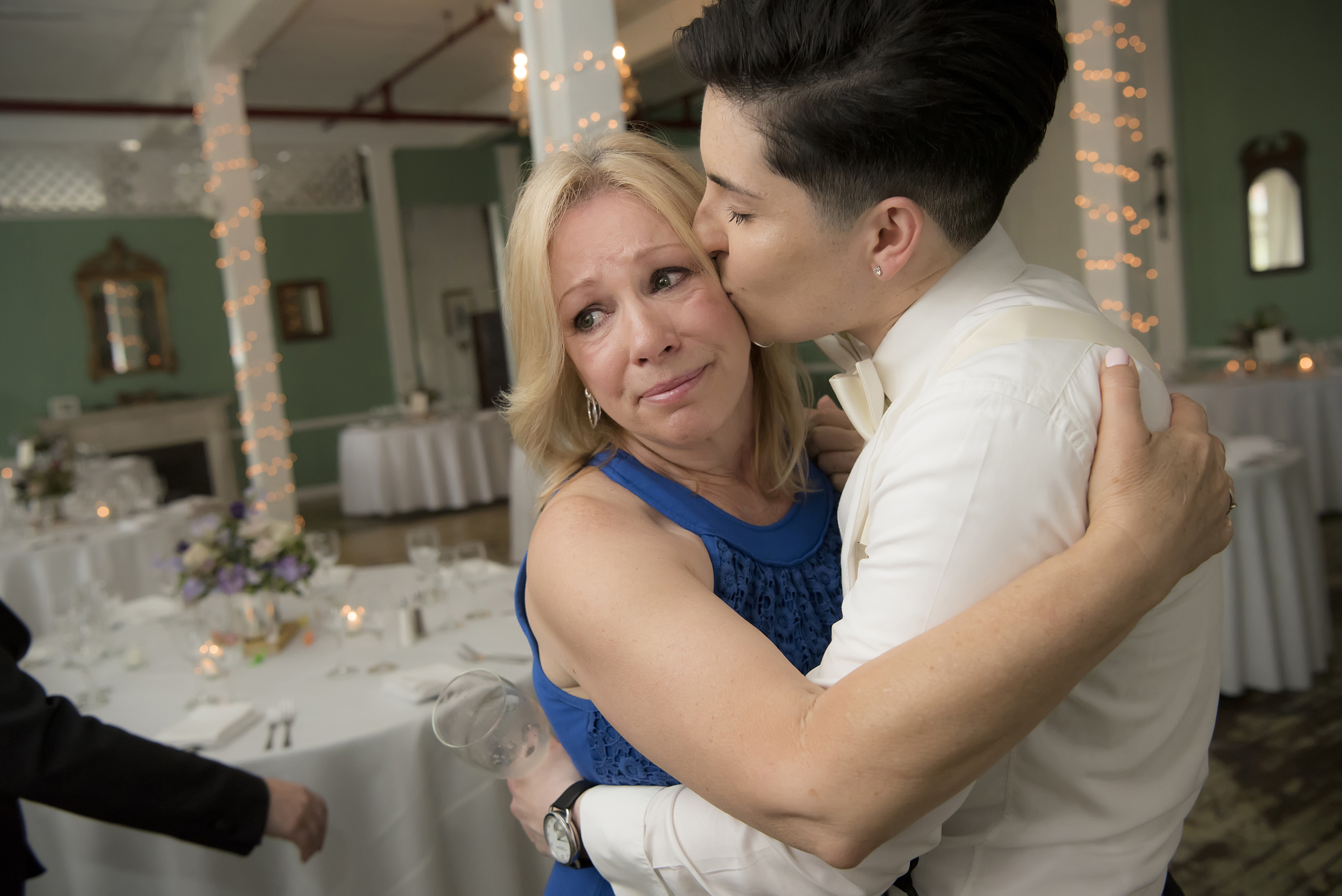 Women tearfully hugs bride with short dark hair wearing a tuxedo shirt, as they stand in an empty reception hall in a photo by Studio A Images