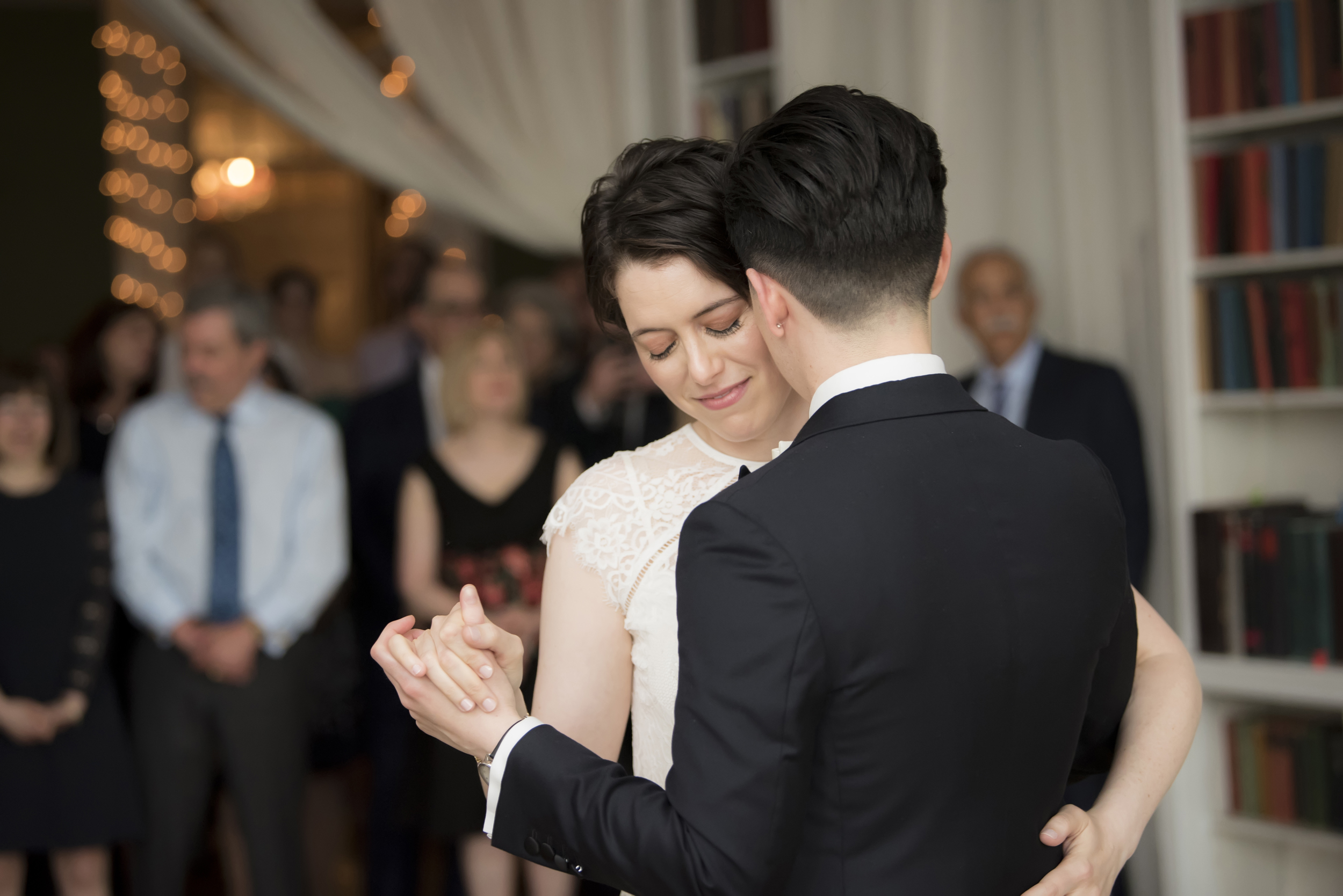 close-up image of a queer wedding couple, one person in a suit and the other in an ivory dress, slow dancing while their guests look on in the background by Studio A Images