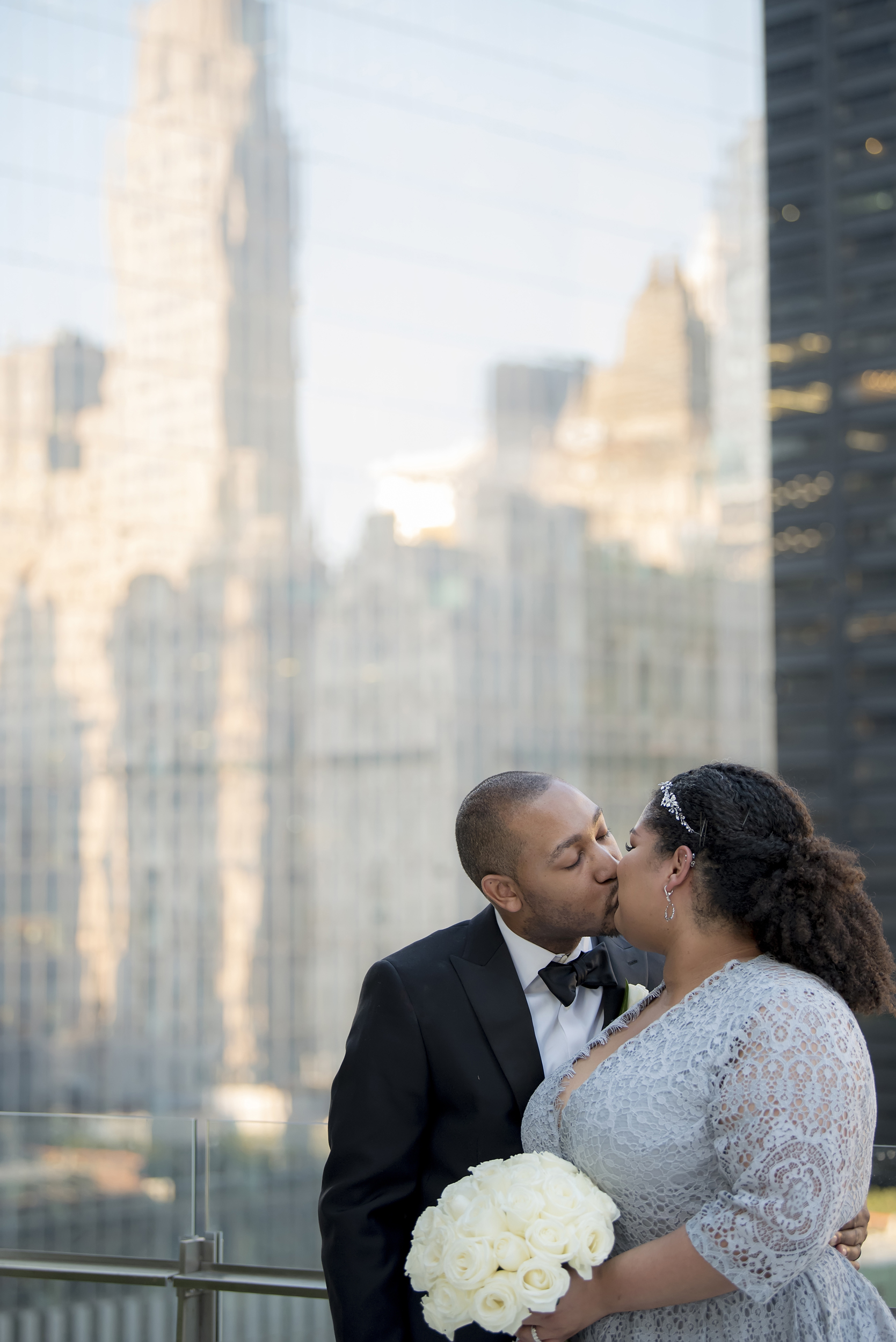 Bride wearing blue lace dress and holding a bouquet of roses kiss on a rooftop with a skyline reflected in the skyscrapers behind them in a photo by Studio A Images