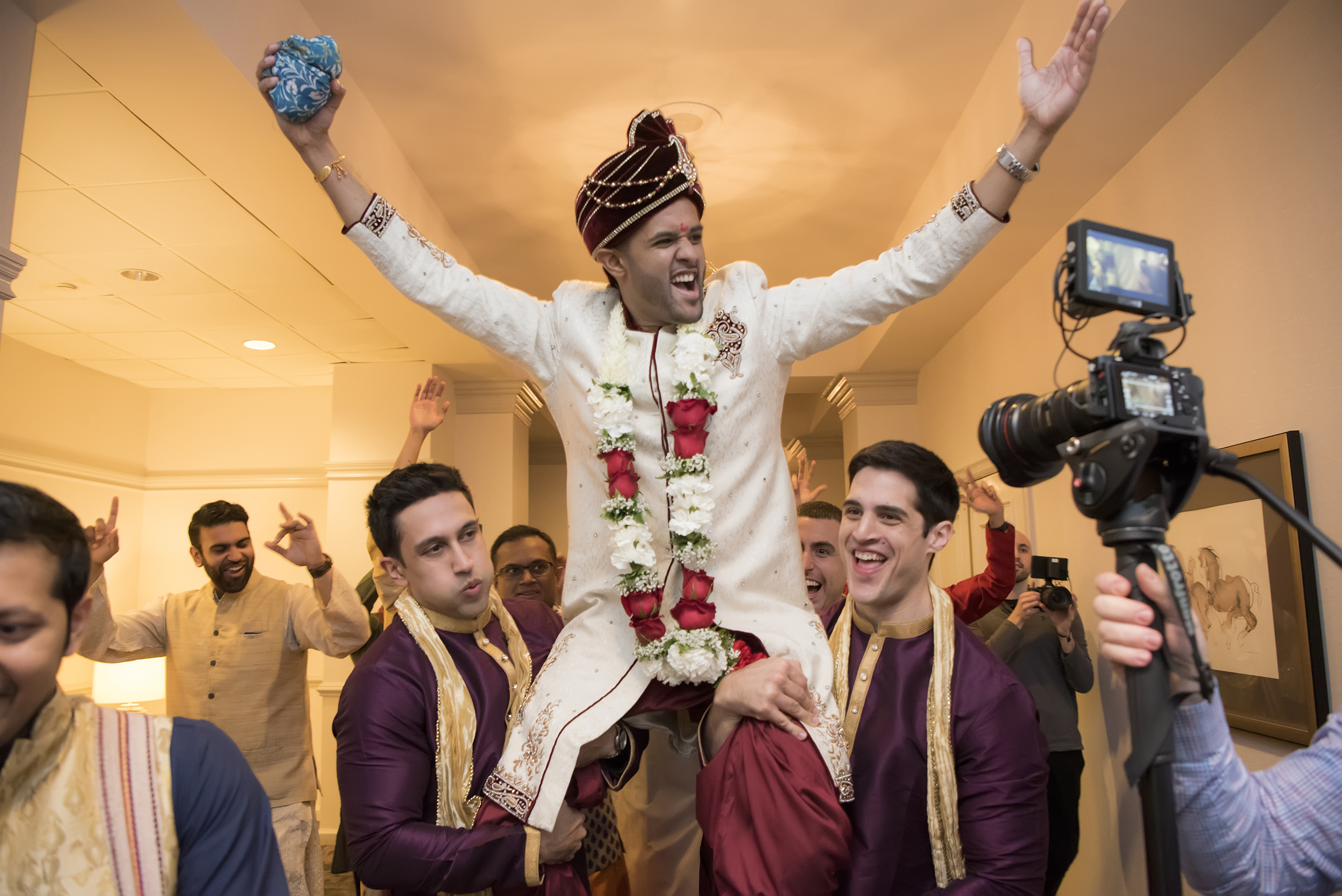 A groom in traditional Indian wedding clothes cheers as he is lifted on the shoulders of two groomsmen, while a videographer records the events in a photo by Studio A Images