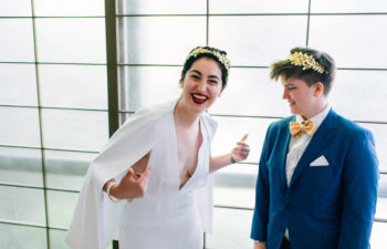 A queer couple in a wedding dress and a suit with bow tie. The person in a wedding dress laughs and points at themself, while the person in a suit smiles and looks on.