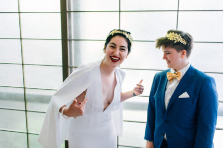 A queer couple in a wedding dress and a suit with bow tie. The person in a wedding dress laughs and points at themself, while the person in a suit smiles and looks on.