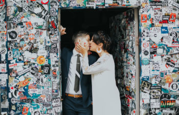 A queer couple kiss in their wedding clothes while standing in a doorway plastered with stickers in a photo by Studio XII Photography