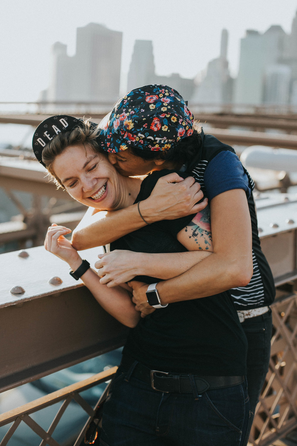 two women embrace in a hug wearing fashion baseball hats covered in flowers and the word "dead" on the brooklyn bridge