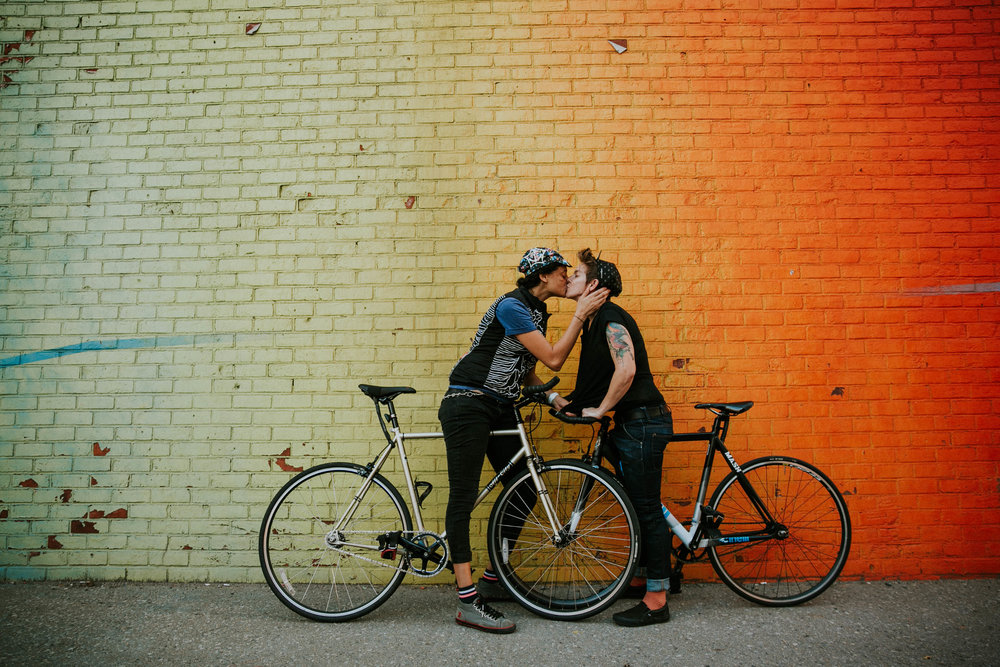 a queer couple pose in front of an ombre wall while standing on bicycles kissing for an LGBTQ engagement session