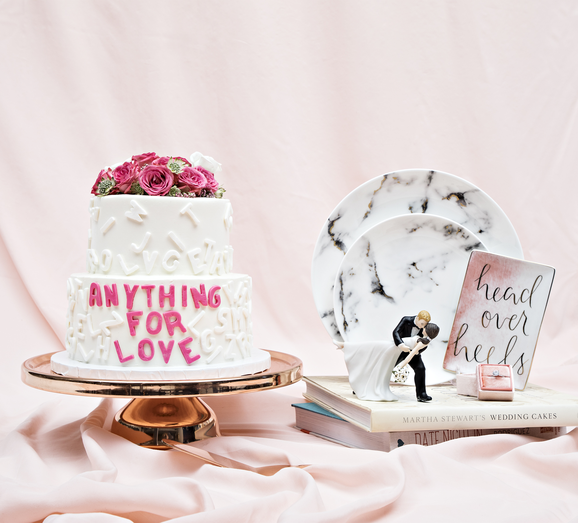 A wedding cake that reads "Anything For Love" next to two marble plates and a cake topper