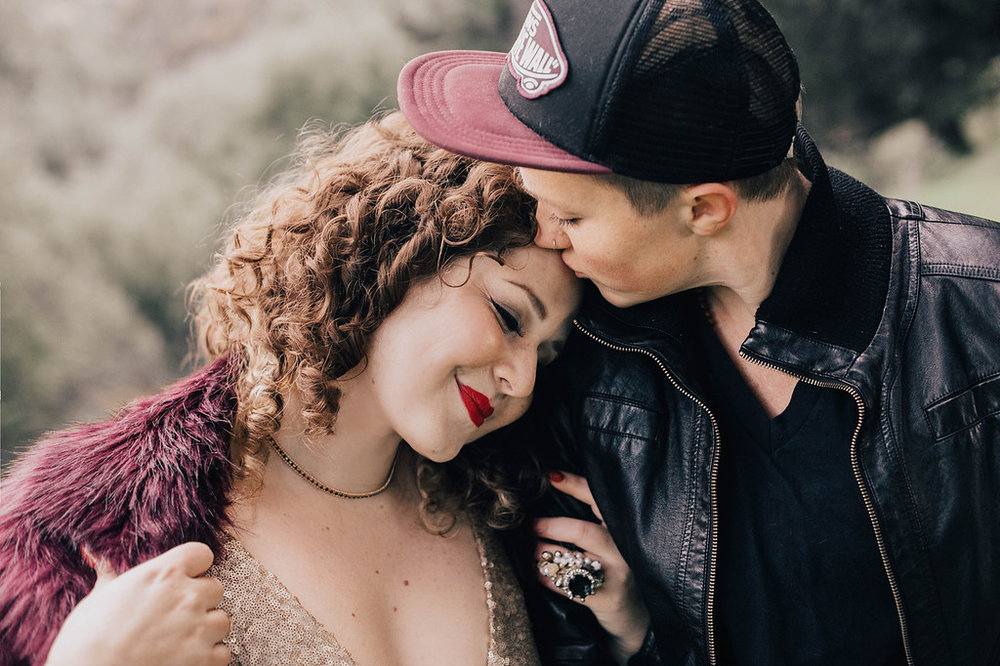 a woman in a fur jacket and sequin dress wearing red lipstick leans against her masculine of center partner in a baseball hat and leather jacket for an engagement shoot