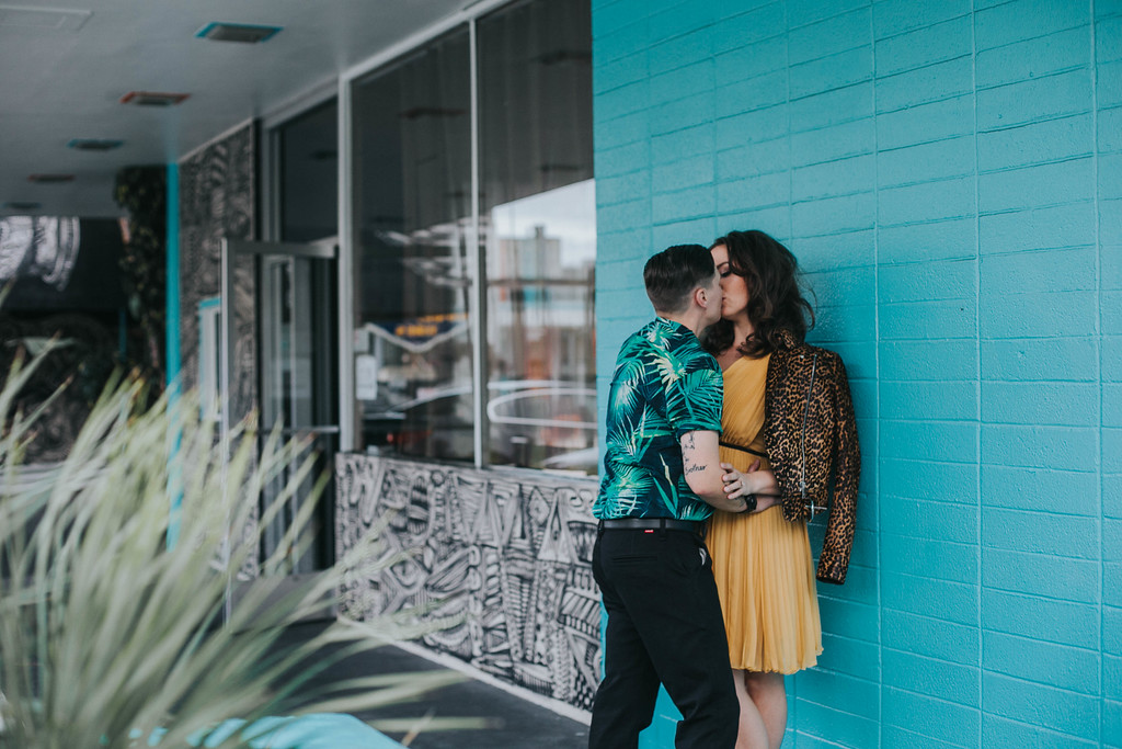 an LGBTQ couple kiss while leaning against a turquoise wall wearing bright patterned clothing for an engagement session