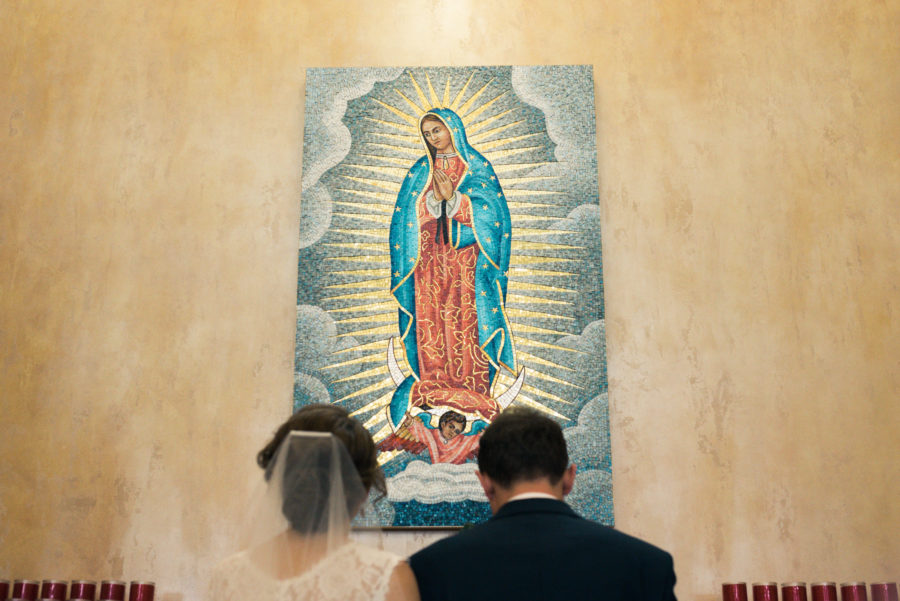 A couple in a wedding dress and a dark suit kneel in prayer in front of a image of Our Lady of Guadalupe