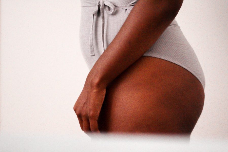A close up portrait of a woman's hip and thigh. She as brown skin and is wearing a light grey bodysuit with a tied waist.