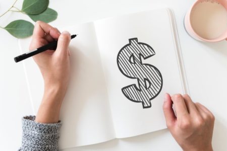 Hands holding a pen over a notebook with a large illustrated dollar sign drawn inside. The notebook sits next to a mug.