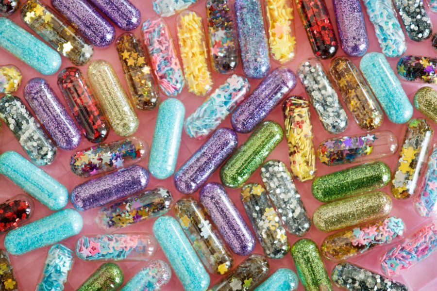 A collection of clear pill capsules filled with different types of brightly colored and metallic glitter on a pink background