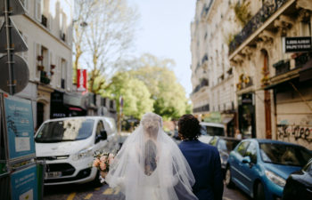A person in a wedding dress and veil walks away from you next to a person in a dark suit on a Paris street with lots of architectural detail