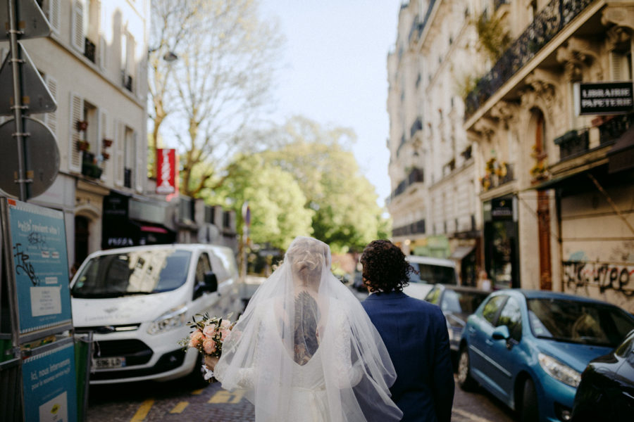 A person in a wedding dress and veil walks away from you next to a person in a dark suit on a Paris street with lots of architectural detail