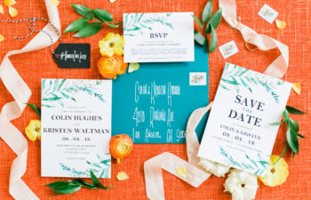 A wedding invitation suite with a tropical botanical theme displayed with ribbon, flowers, and foliage on an orange background