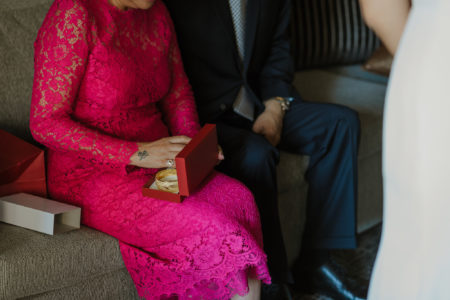 A woman in a bright pink lace dress opens a red jewelry gift box in her lap containing bracelets